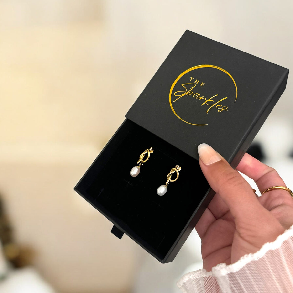 A pair of pearl drop earrings presented in a black box with 'The Sparkles' logo, held by a person. The earrings have gold-colored accents and a pearl drop.