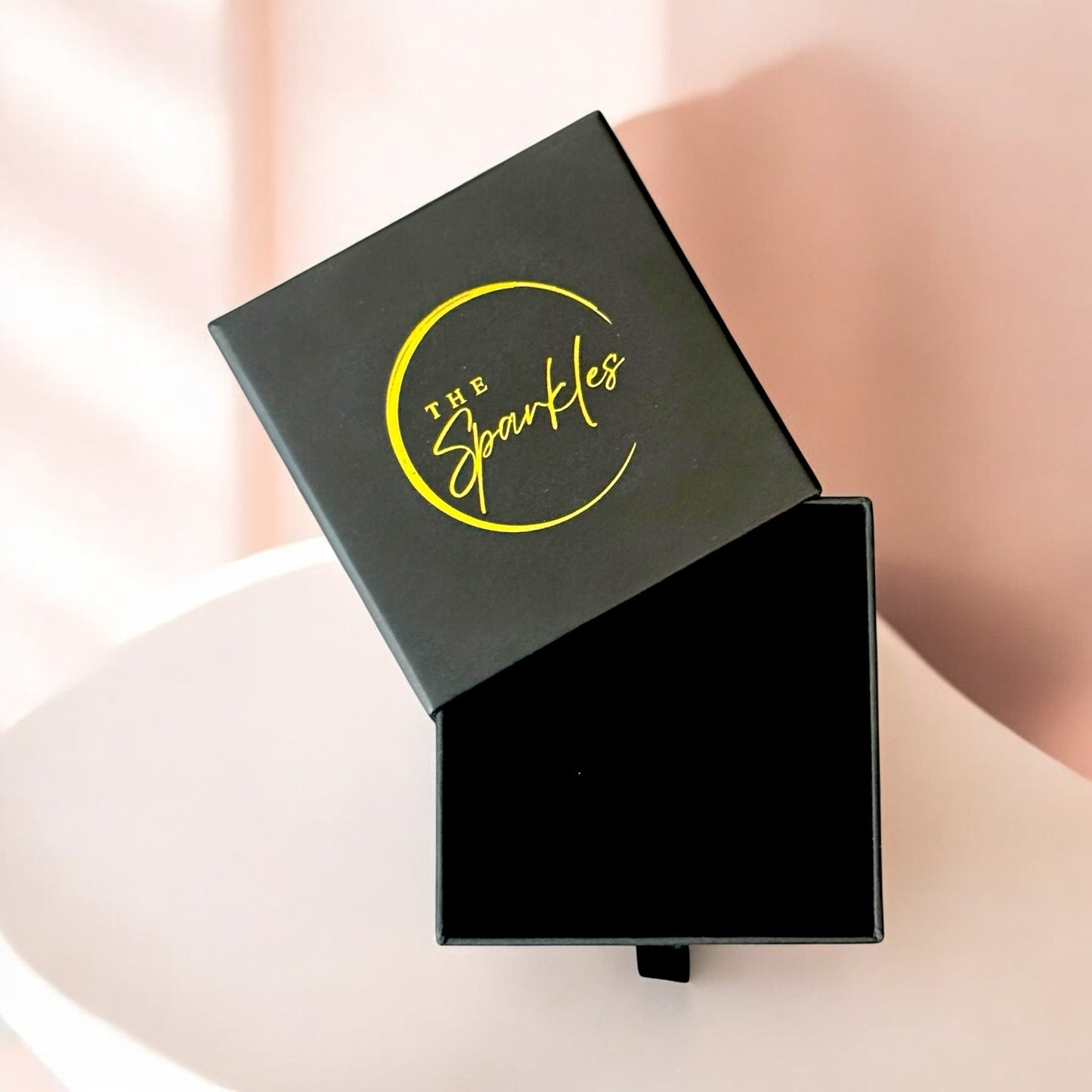 A black gift box with a yellow logo on the lid, labeled 'the sparkles', presented against a light pink background.
