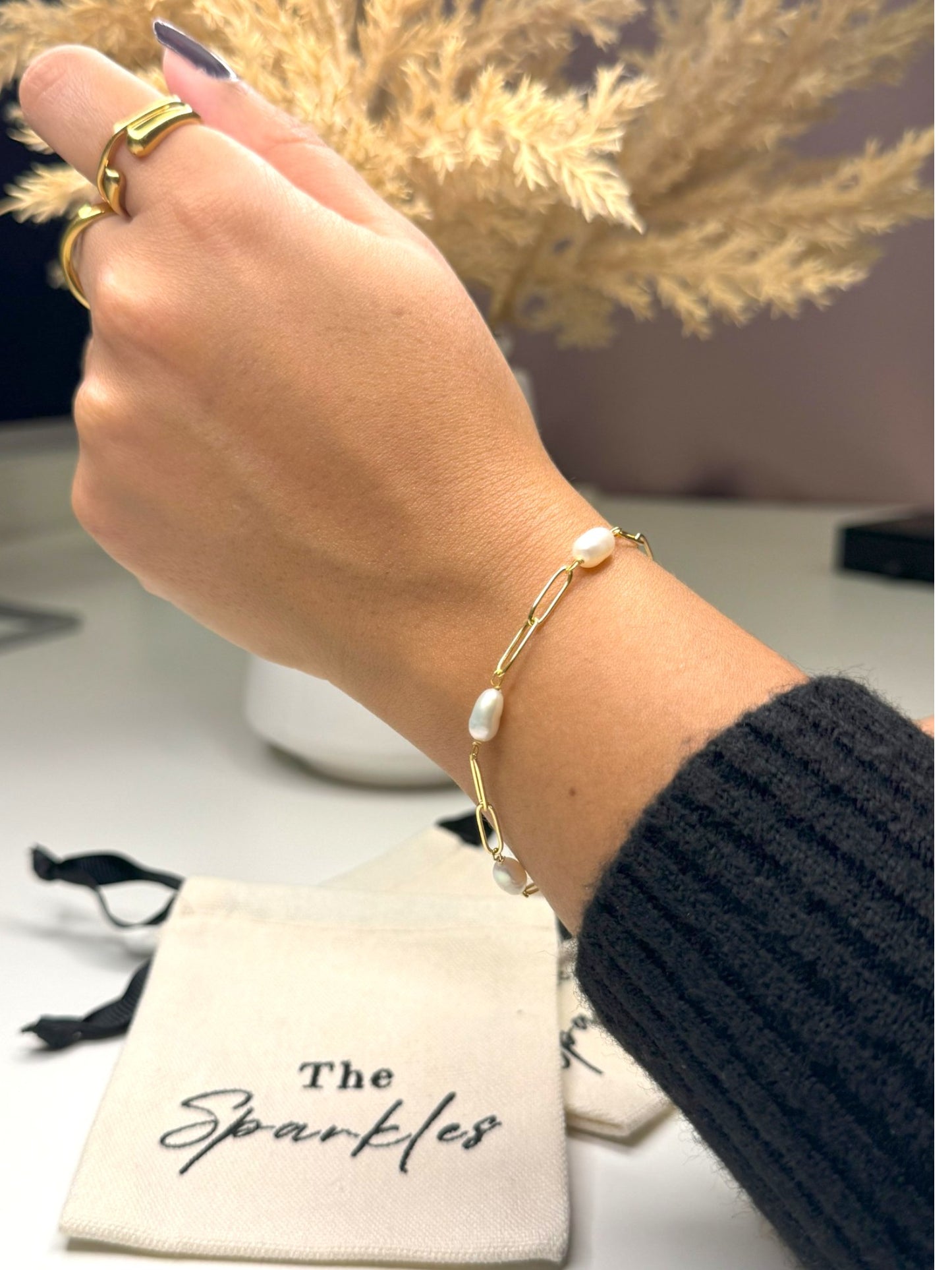 A person's wrist wearing a sterling silver bracelet with 14K gold plating and freshwater pearls, alongside a white drawstring pouch with 'The Sparkles' branding.