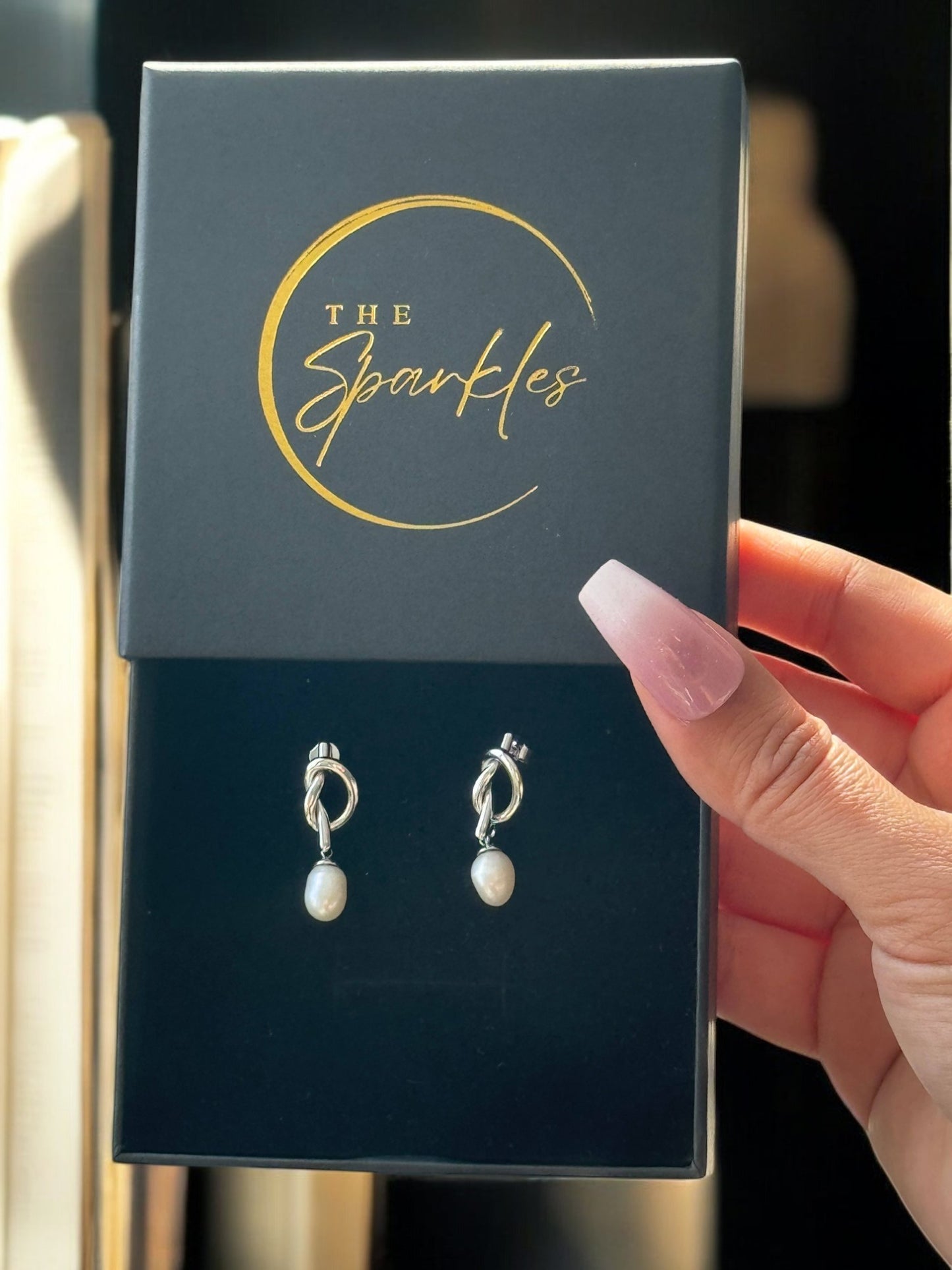 A pair of pearl drop earrings presented in a black box with 'The Sparkles' branding, held by a person.