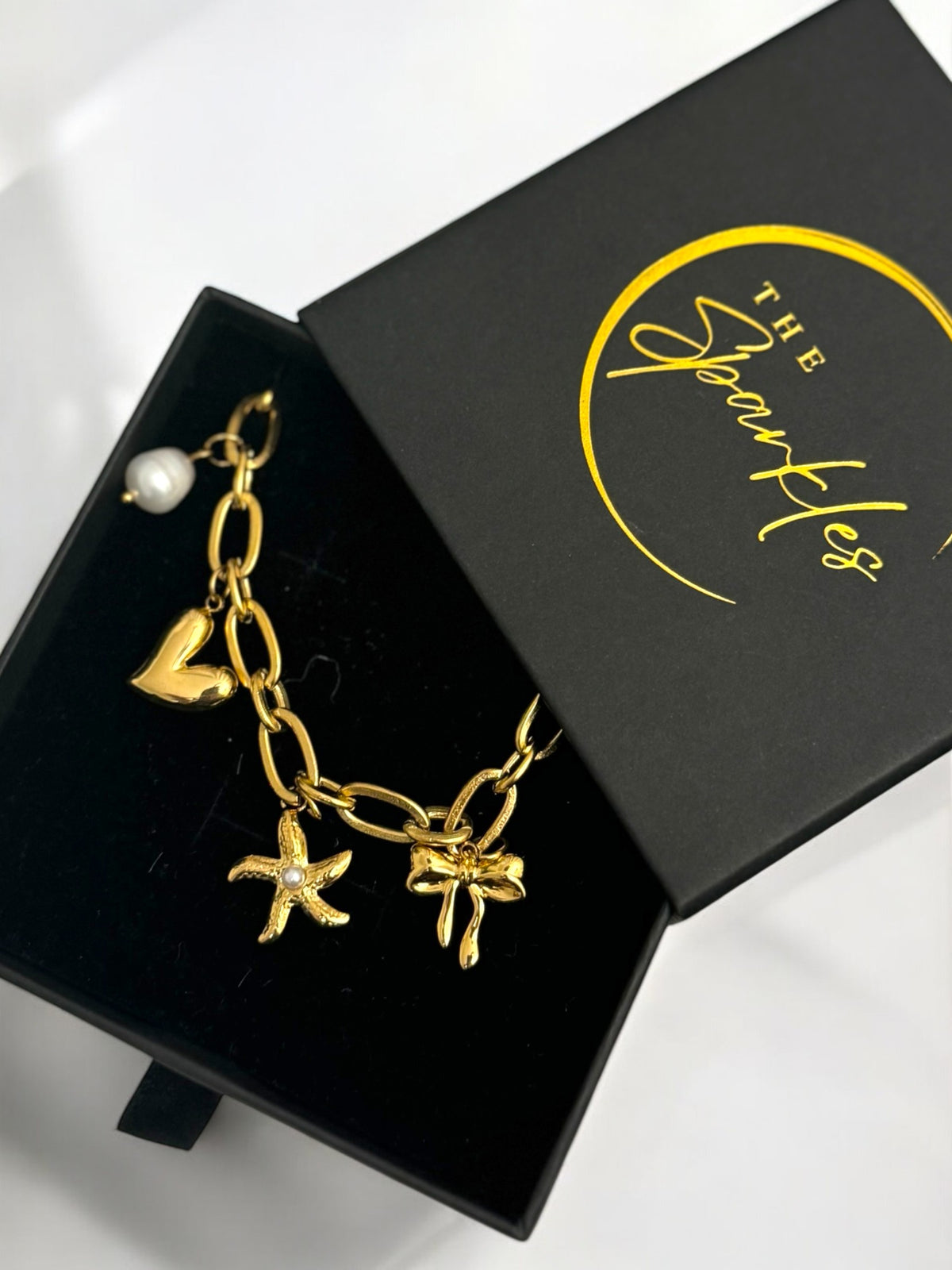 A gold-plated paperclip chain necklace with various charms displayed inside a black box with gold accents, branded as 'The Sparkle'.