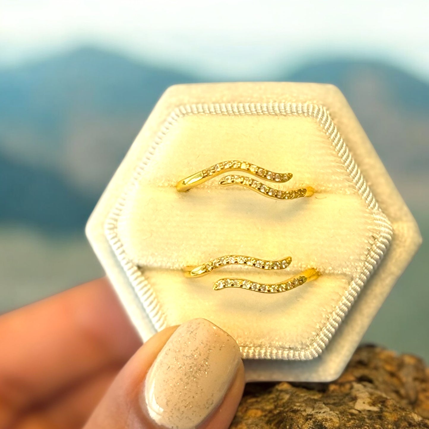 Two gold-colored rings with a geometric design, displayed on a white surface with a mountainous background.