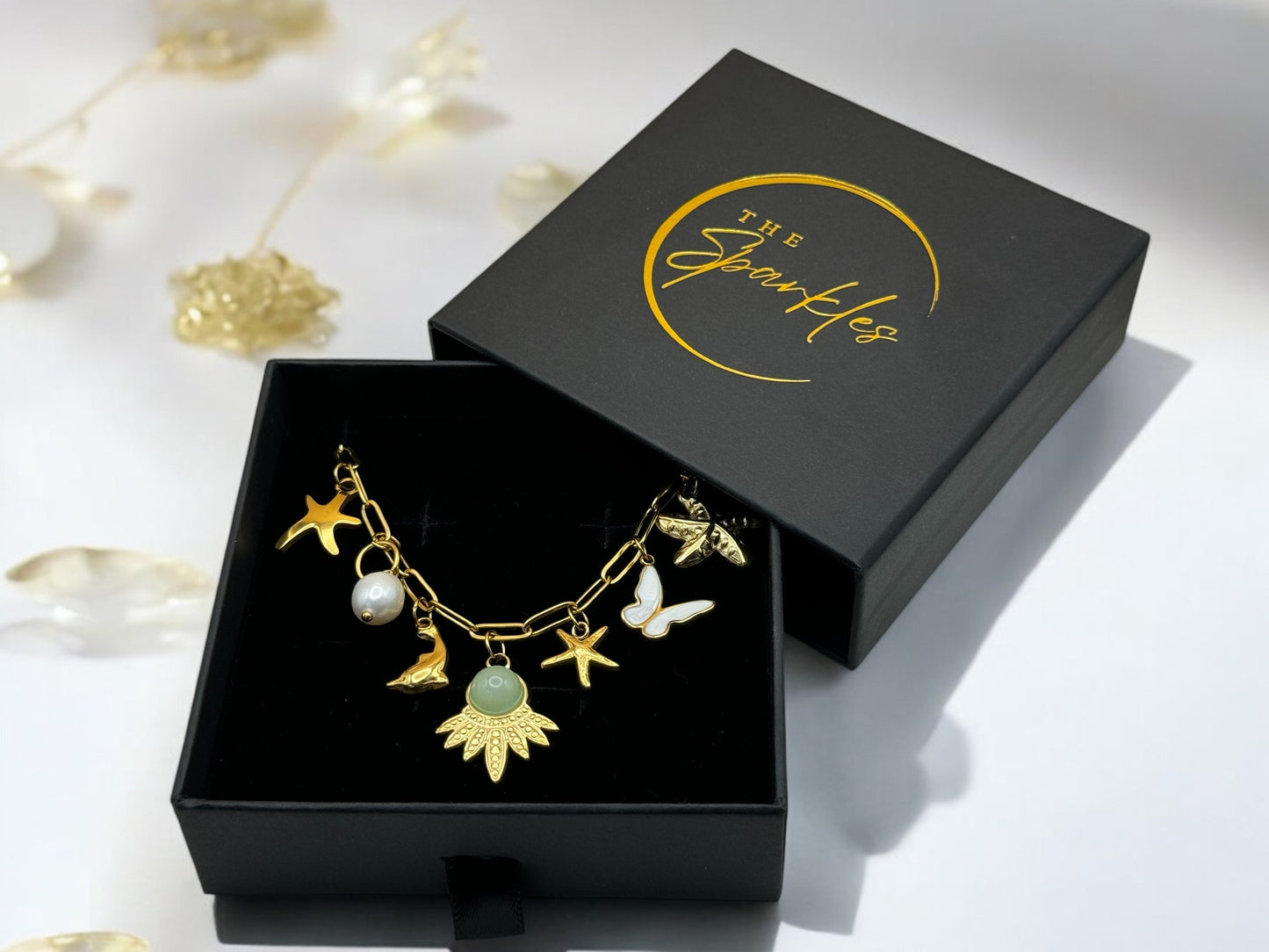 A personalized 18K gold plated necklace with various charms including a butterfly, star, and leaf, displayed in an open black box with 'The Sparkles' branding.