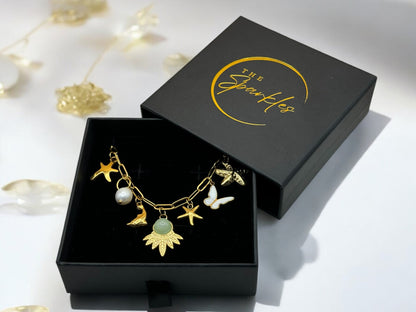 A personalized 18K gold plated necklace with various charms including a butterfly, star, and leaf, displayed in an open black box with 'The Sparkles' branding.